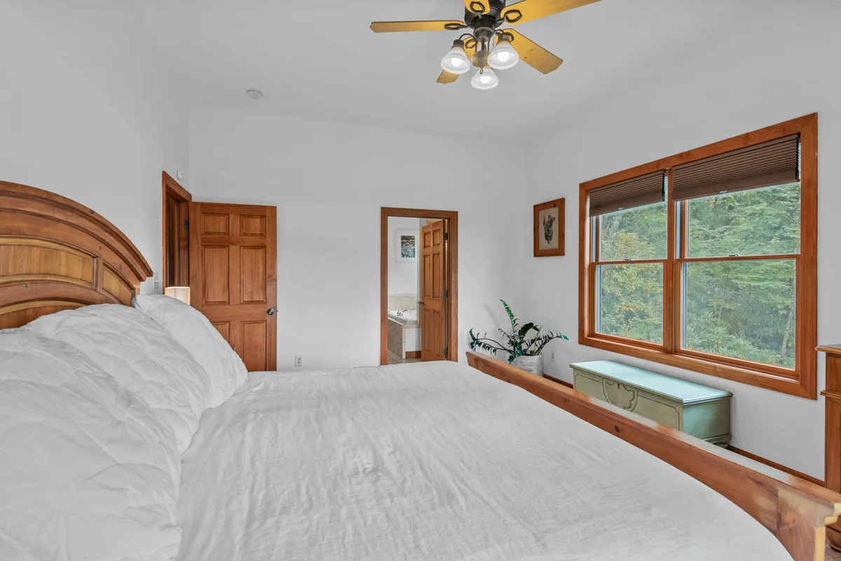 A serene primary bedroom featuring a king-sized bed with crisp white bedding, warm wooden furniture, and a window inviting morning light and green outdoor views. The ceiling fan and uncluttered décor create a restful, airy atmosphere ideal for relaxation.