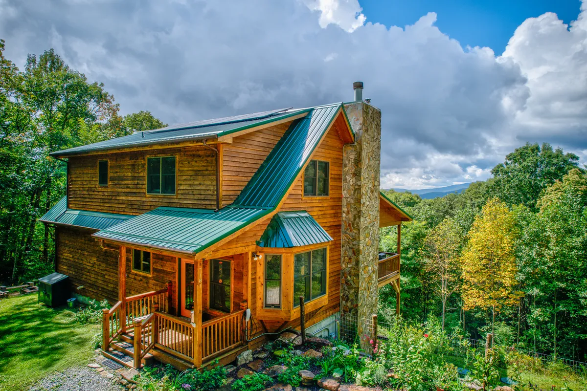 The rustic wooden exterior of Peaceful View is nestled among tall trees, with a green metal roof and stone chimney. The home exudes seclusion and is surrounded by nature, hinting at the sense of privacy and adventure the location promises year-round.