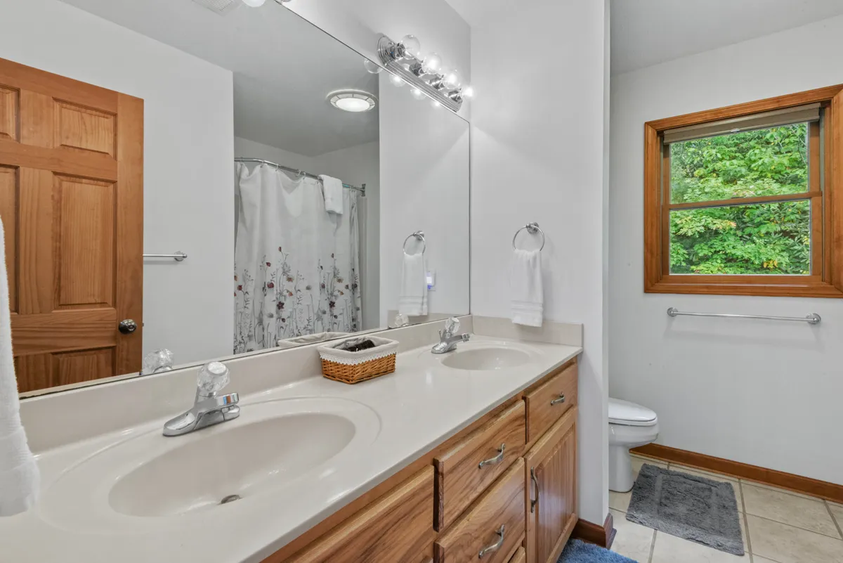 A fresh, serene bathroom with a double sink vanity, wooden cabinetry, a large mirror, and a window offering views of greenery. The space is light-filled and designed for comfort, serving as a convenient place for guests to refresh and recharge between games.