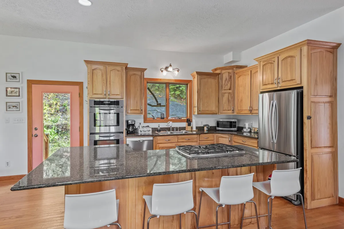 A close view of the kitchen, with a granite-topped island, stainless steel appliances, custom wooden cabinetry, and a window view. The space is set up for cooking and casual breakfasts, exuding warmth and functionality.
