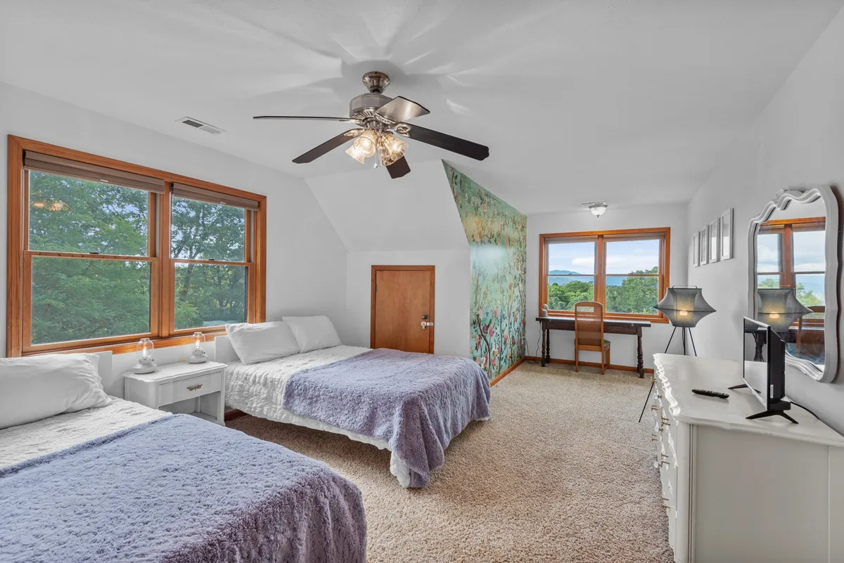 A bright upstairs bedroom with two cozy beds dressed in soft lavender bedding, large windows framing the scenic outdoors, and a small desk and chair for work or play. Relaxed artwork and décor add to the welcoming family atmosphere.