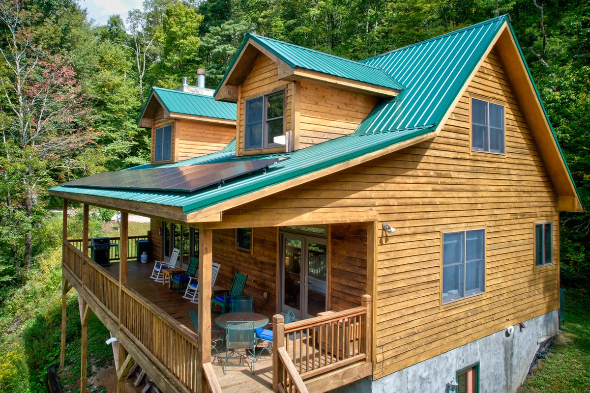 This image shows the exterior of the home, featuring a welcoming wooden deck with seating surrounded by lush trees. The green metal roof and the outdoor seating area suggest a space perfect for gathering after a day of adventure. The deck area is shown as an inviting place for socializing or relaxing in nature.