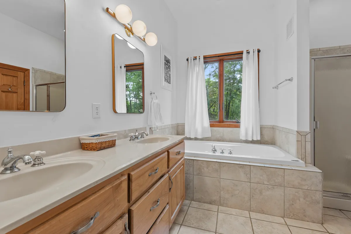A spa-like ensuite bathroom with a large soaking tub positioned beneath a window overlooking lush greenery, a double sink vanity with ample counter space, and elegant lighting. The room exudes tranquility and luxury.