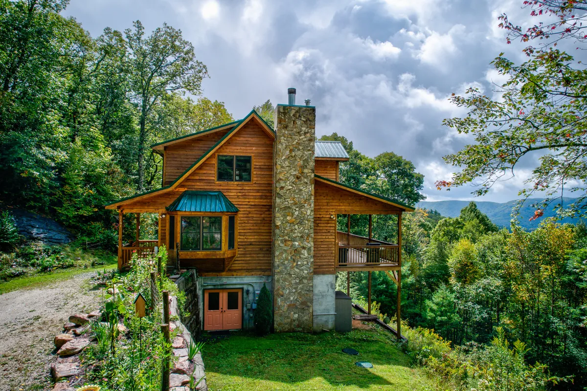 The hero image showcases a charming wooden mountain home with a striking stone chimney set against a lush backdrop of greenery and expansive mountain views. The covered deck invites relaxation, while the tranquil setting hints at both sunrise coffee moments and cozy evenings under the stars.