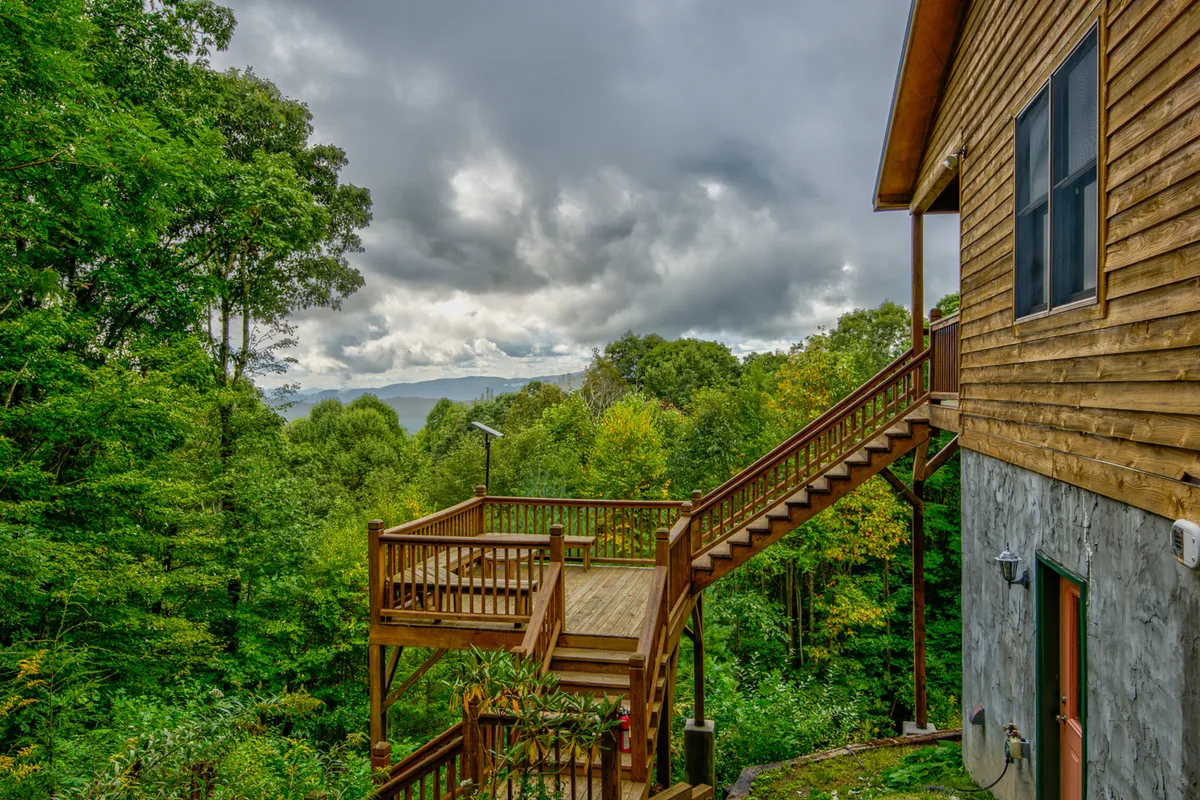 A serene wooden deck overlooks distant mountains, surrounded by lush trees and nature. The inviting deck space, complete with seating, embodies a place to relax after a day of adventure or gather with friends and family while taking in panoramic mountain views.