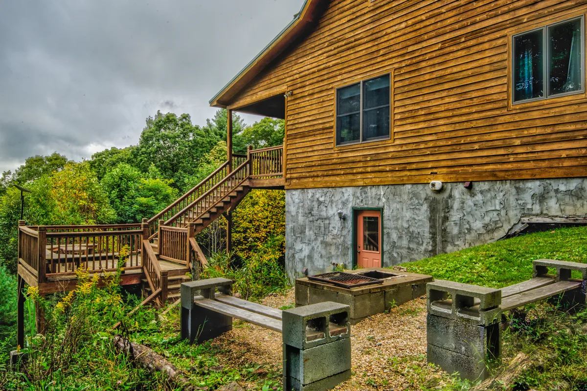 This image features a spacious wooden deck with a staircase leading down to a fire pit area, surrounded by lush greenery. The setup invites guests to relax outdoors, enjoy barbecues, and gather around the fire as the sun sets.