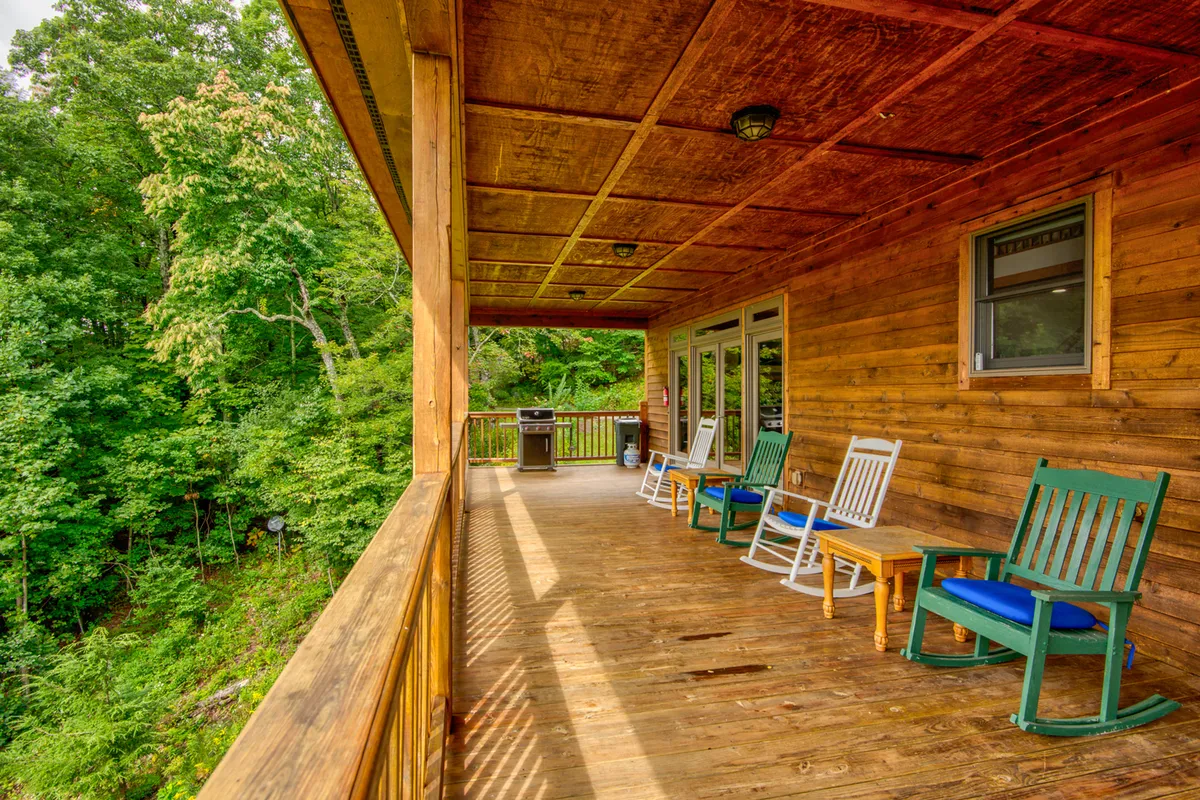 A serene exterior view showing a covered deck with rocking chairs, a wooden railing, a propane grill, and lush greenery in the background. The setup is perfect for sipping coffee or enjoying a mountain BBQ while taking in the peaceful surroundings.